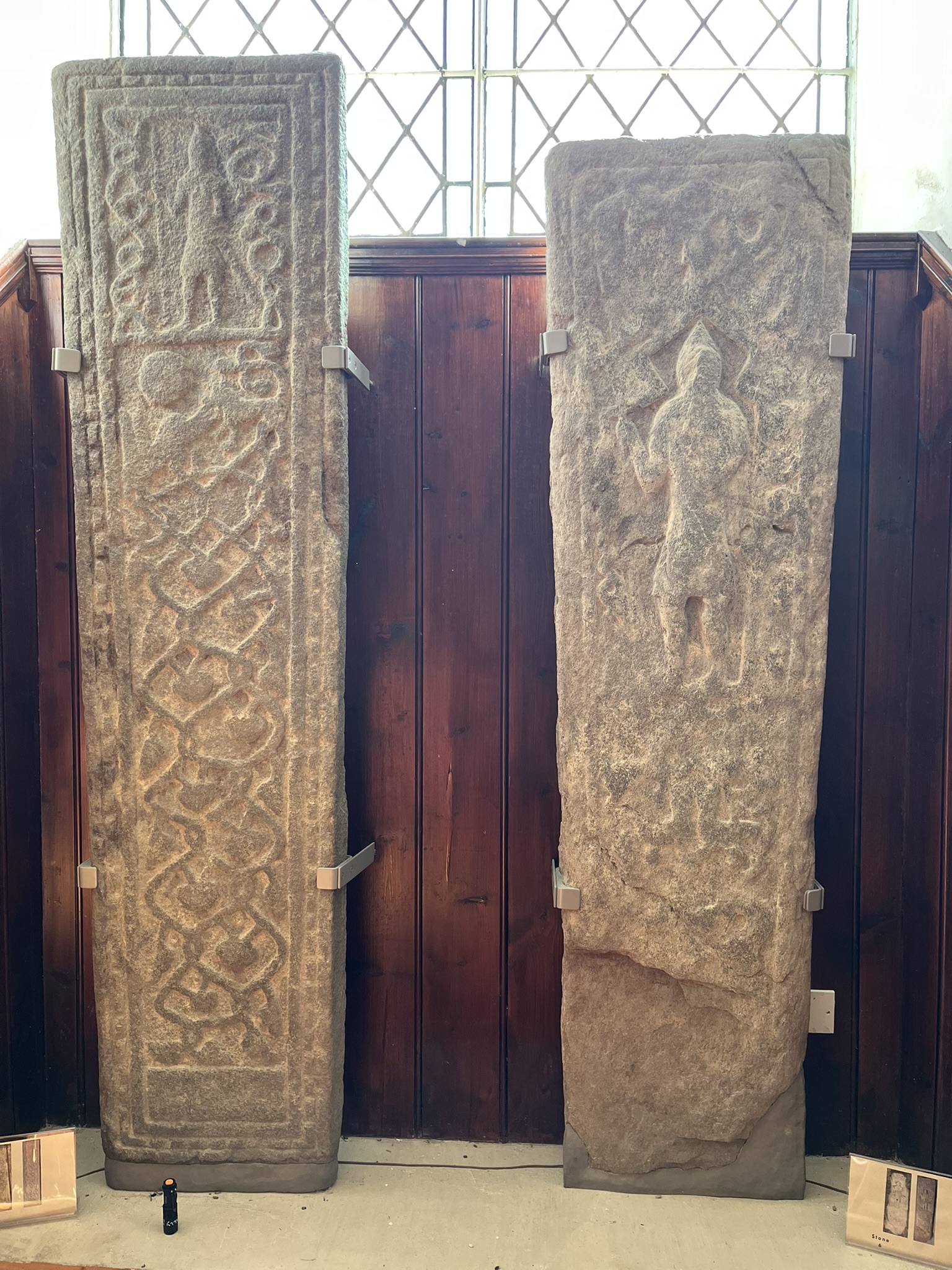 A pair of slabs displayed against the panelling: at left, deep-cut bands of interlace below a worn figure; at right, a tall standing warrior.