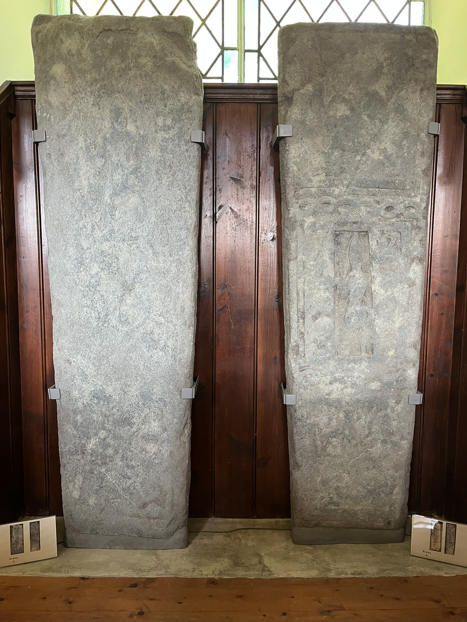 Two of the more heavily weathered slabs standing against the panelling, their carving worn nearly smooth by long exposure.