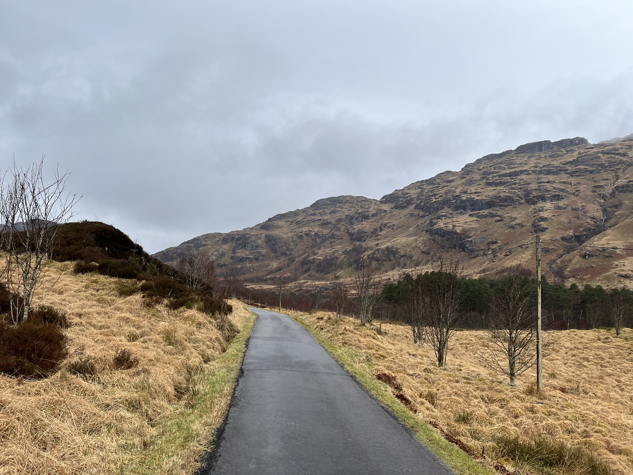 A single-track tarmac road climbing gently away into the mouth of Glen Gyle between rough grass verges, with the slopes of the surrounding hills rising ahead under an overcast sky.