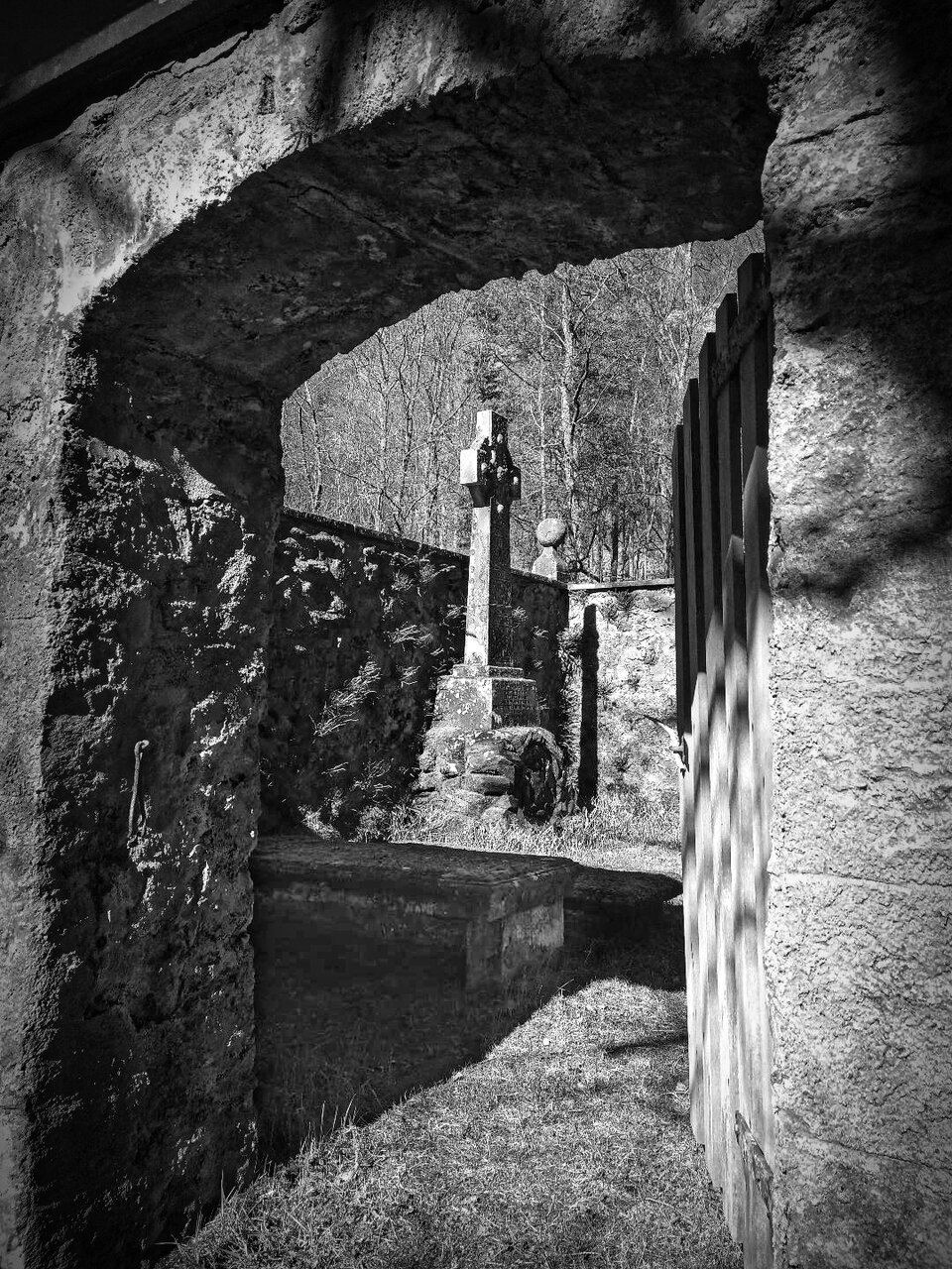 Black and white photograph looking through the stone archway of the burial enclosure to a tall cross-headed memorial standing inside against the back wall, with bare trees beyond.