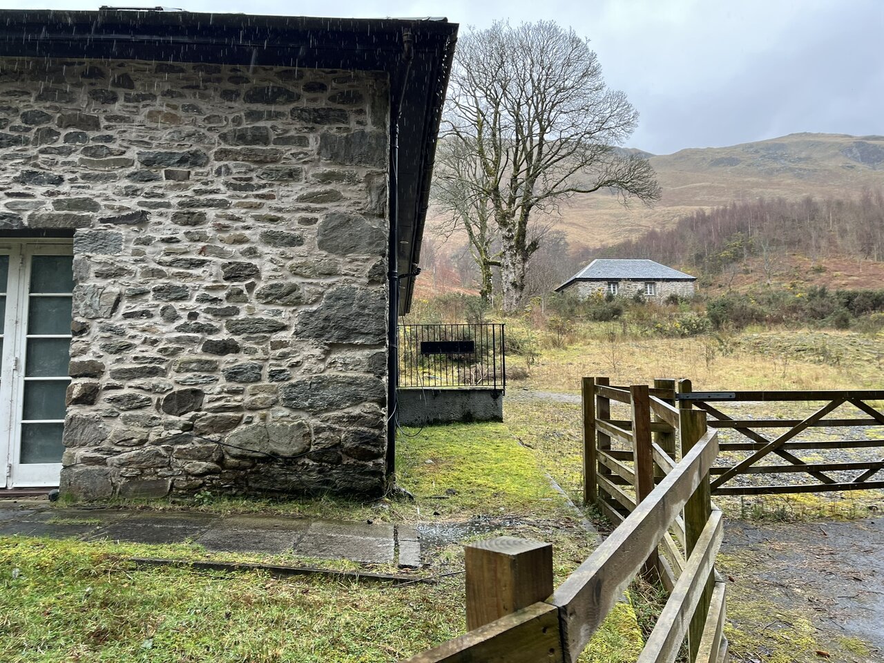 A small stone bothy with a grey roof standing in a field below the hillside at Glengyle, seen past the gable corner of a rubble building and a wooden field gate, with rain falling.