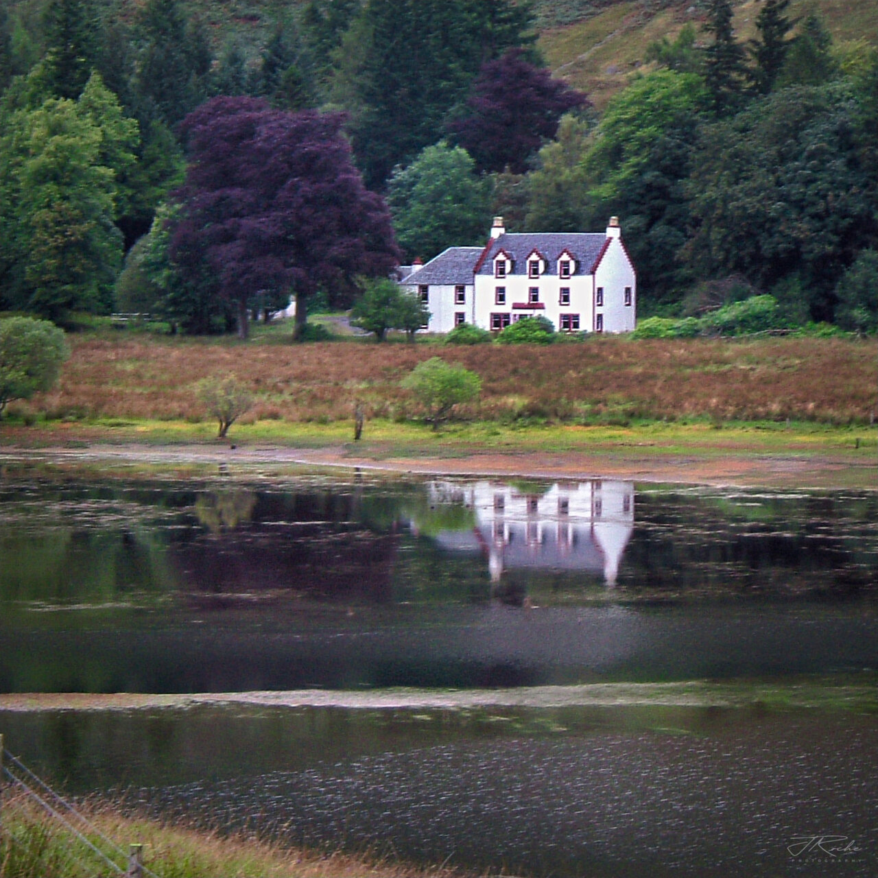 Glengyle House seen from across the loch, the white house set among trees on the shore at the foot of the hillside and reflected in the still water in front of it.