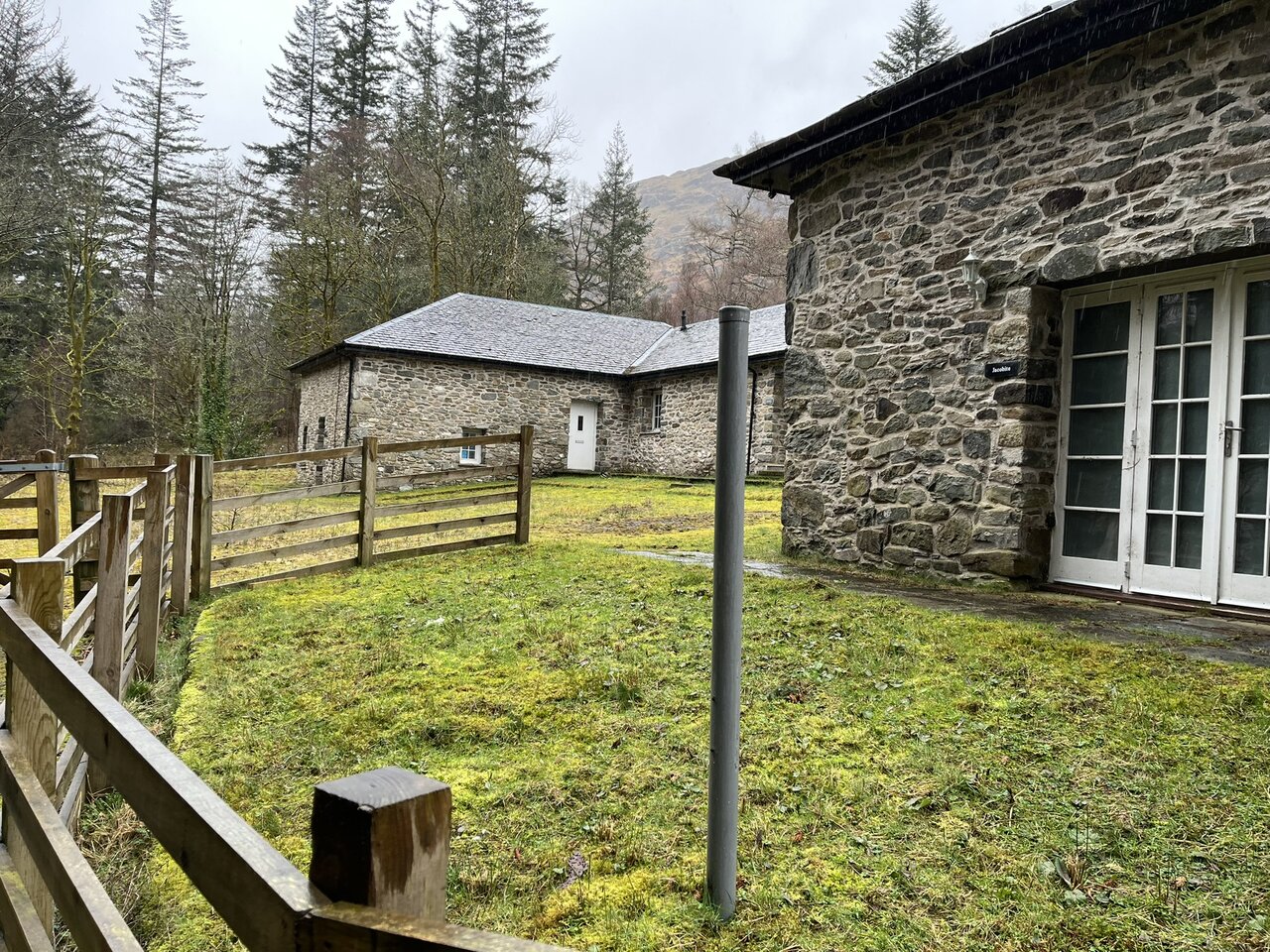 The steading at Glengyle House - a long stone range with a grey slate roof set behind a post-and-rail fence, with the corner of a second rubble-built block carrying white-painted French doors in the foreground and a wooded hillside behind.