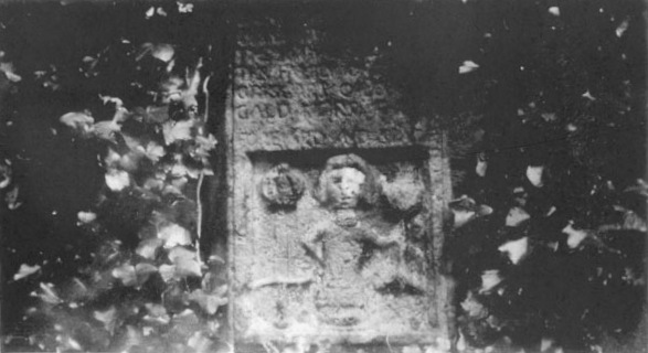 Vintage black and white photograph of the weathered stone relief on the enclosure wall, an inscribed panel above a carved central figure flanked by smaller emblems, framed by overhanging foliage.