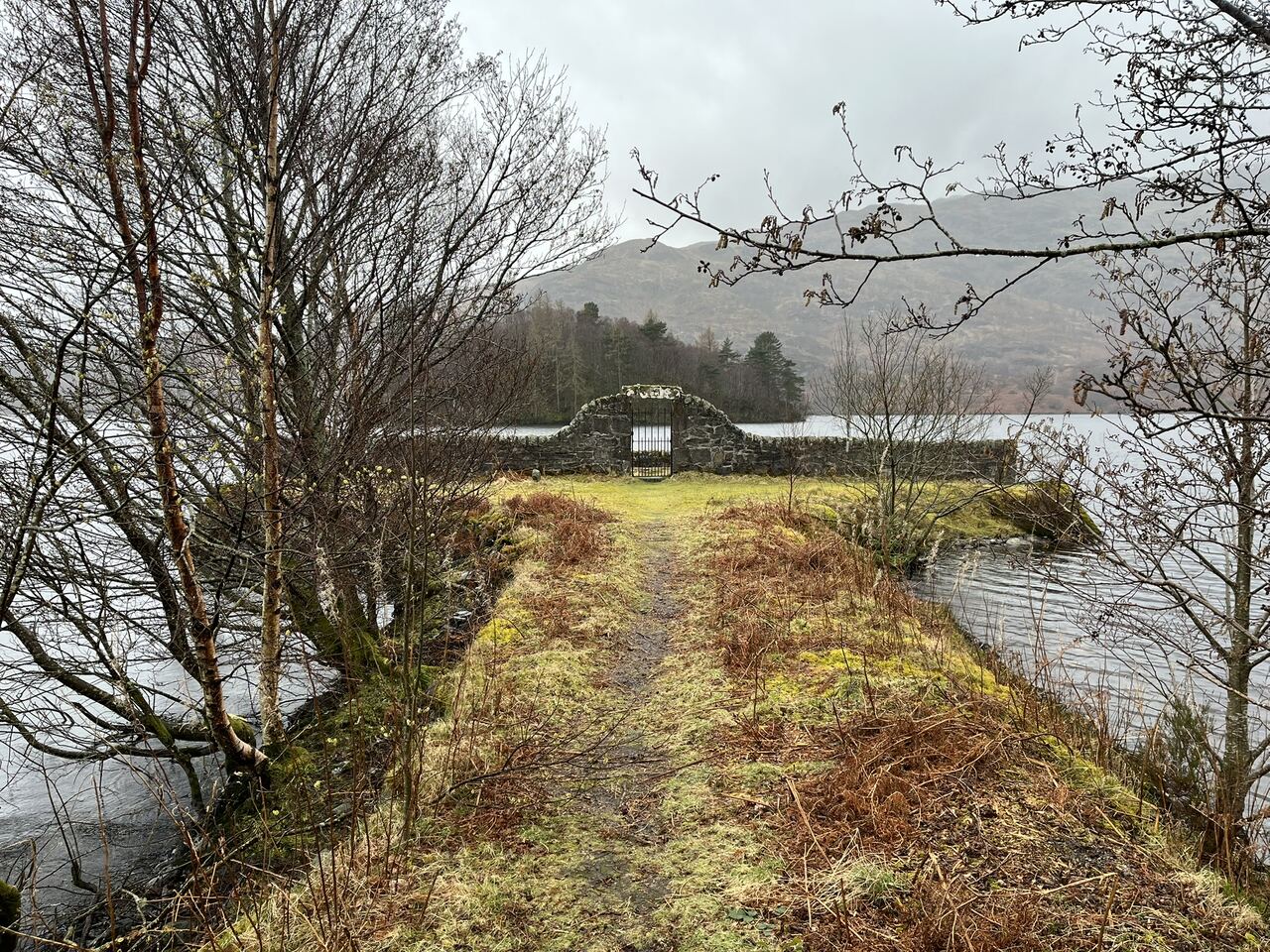 Looking down the grassy path between trees to the stone entrance of the Portnellan burial enclosure, with Loch Katrine and the hills beyond.