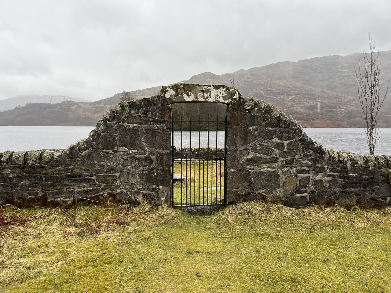 The stone entrance arch with its iron gate, seen head on from outside, looking through into the grassy enclosure with Loch Katrine and the hills beyond.