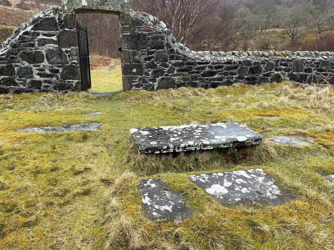 Inside the inner enclosure, looking back towards the gated stone entrance, with a raised slab in the centre and lichen-covered flat slabs set in the turf around it.
