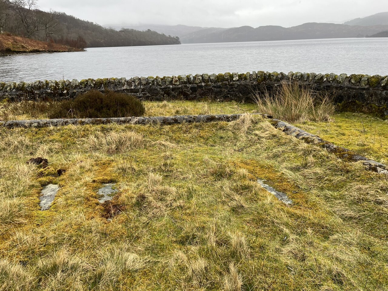 The low grass-covered foundations of the outer enclosure with recumbent slabs, the lochside wall, and Loch Katrine beyond.