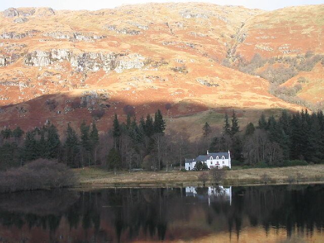 Glengyle House at the head of Loch Katrine - a white two-storey cottage standing among trees at the foot of a richly-coloured hillside, reflected in the loch in front of it.